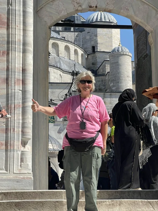 A person in a pink shirt stands smiling, giving a thumbs-up in front of a historic mosque with domes and arches.