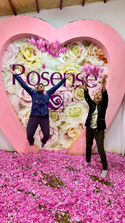 Two people joyfully toss pink petals in the air, standing in front of a large heart-shaped display filled with roses.