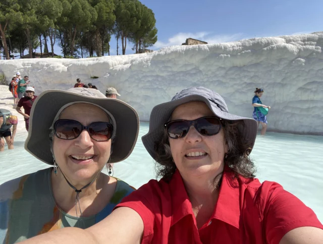 Two people wearing hats take a selfie in front of travertine terraces with several other visitors visible in the background.