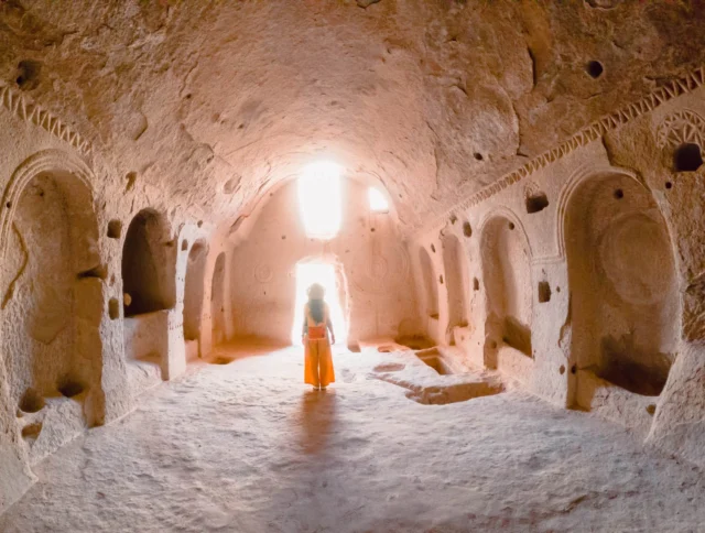 Person in orange clothing stands in a cave-like room with intricate carvings and niches, illuminated by sunlight streaming through the entrance.