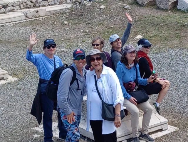 A group of seven people posing outdoors, some waving, on a stone structure. They are dressed casually and wearing caps. Grass and stone fragments are visible in the background.