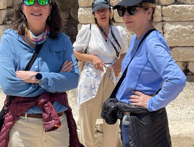 Three people in casual clothing and hats stand in front of ancient stone ruins, with two posing for the camera.