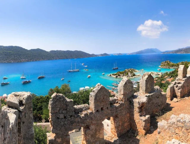 View from stone ruins overlooking blue sea with sailboats and green hills in the background under a clear sky.