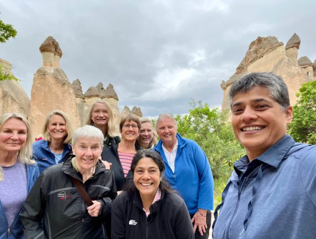 A group of nine people smiling for a selfie in front of unique rock formations under an overcast sky.