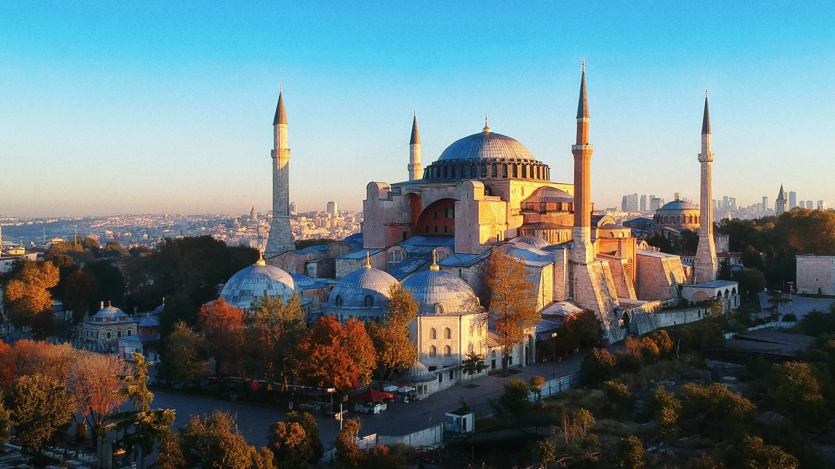 Aerial view of the Hagia Sophia in Istanbul, showing its large dome, four minarets, and surrounding trees with a cityscape backdrop under a clear sky.