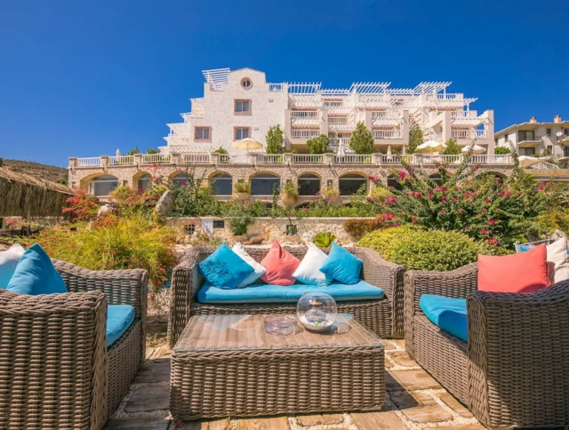 Outdoor patio with wicker furniture and colorful cushions in front of a large white building with terraces and greenery. Clear blue sky in the background.