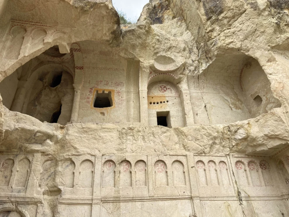 Ancient carved rock face with arched niches and openings, featuring faded red patterns and weathered surfaces.