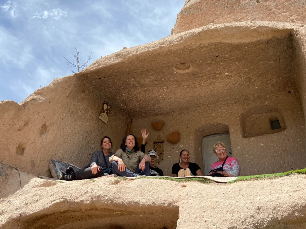 Five people smiling and sitting at the entrance of a cave-like structure carved in rock against a blue sky.