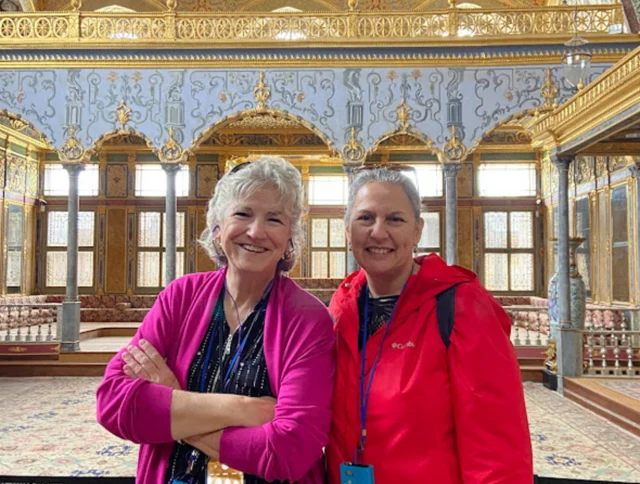Two people standing in an ornate room with decorative ceilings and large windows. They are wearing lanyards and smiling.