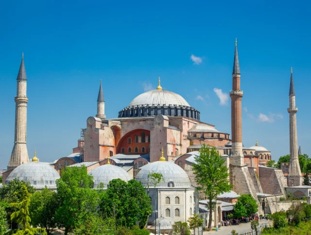 Exterior view of Hagia Sophia, featuring its grand dome and four minarets under a clear blue sky, surrounded by greenery.