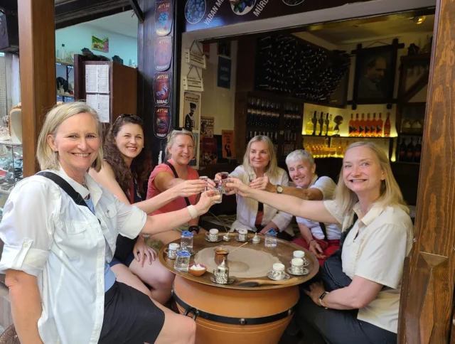 Six people sit around a round table in a cozy cafe, clinking glasses and smiling. Coffee cups and a small tray are on the table. Wine bottles and posters decorate the background.