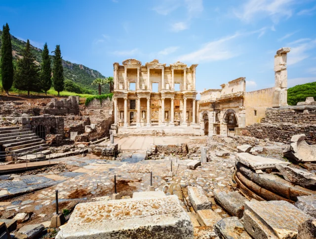 Ancient ruins of the Library of Celsus in Ephesus, Turkey, with stone columns, steps, and surrounding debris under a blue sky.