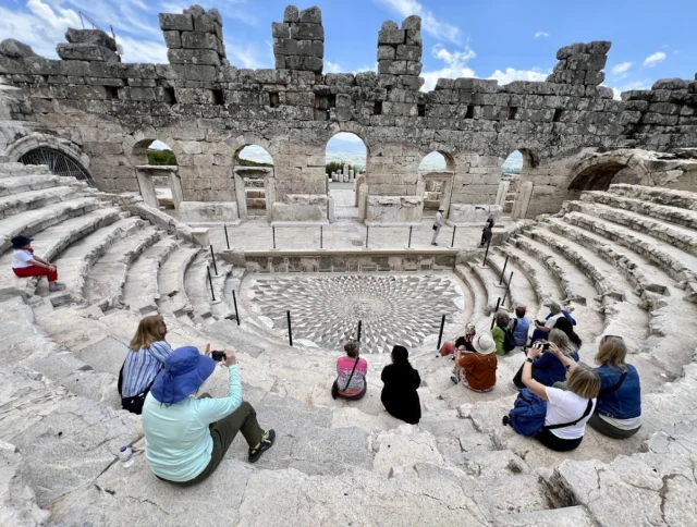 People sitting and taking photos on ancient stone steps in a historic amphitheater with weathered arches under a partly cloudy sky.