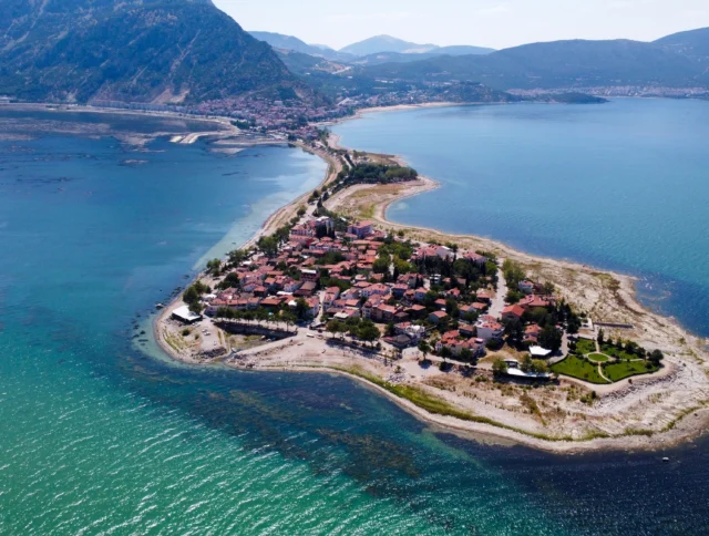 Aerial view of a small peninsula with houses and greenery, surrounded by blue water and mountains in the background.