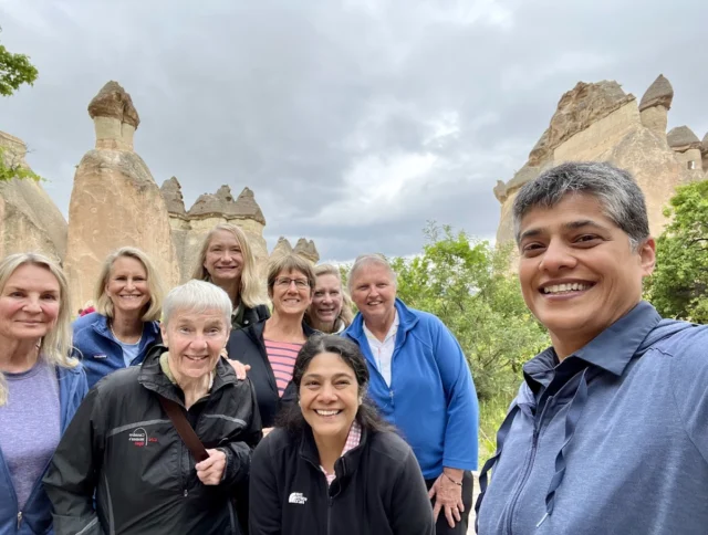 A group of nine people stand smiling in front of unique rock formations under a cloudy sky.