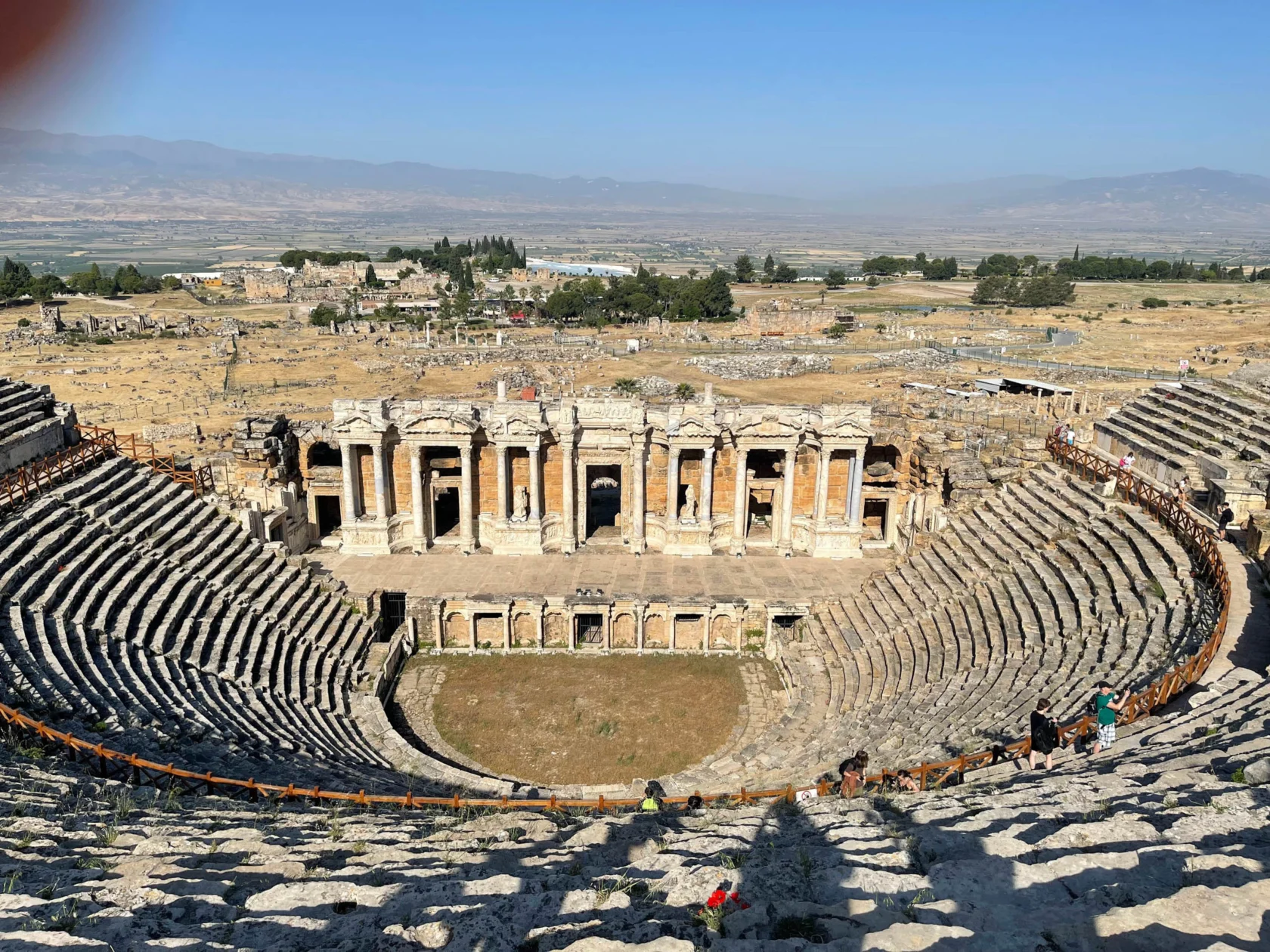 Ancient stone amphitheater with rows of seating, a central stage, and surrounding ruins under a clear sky.