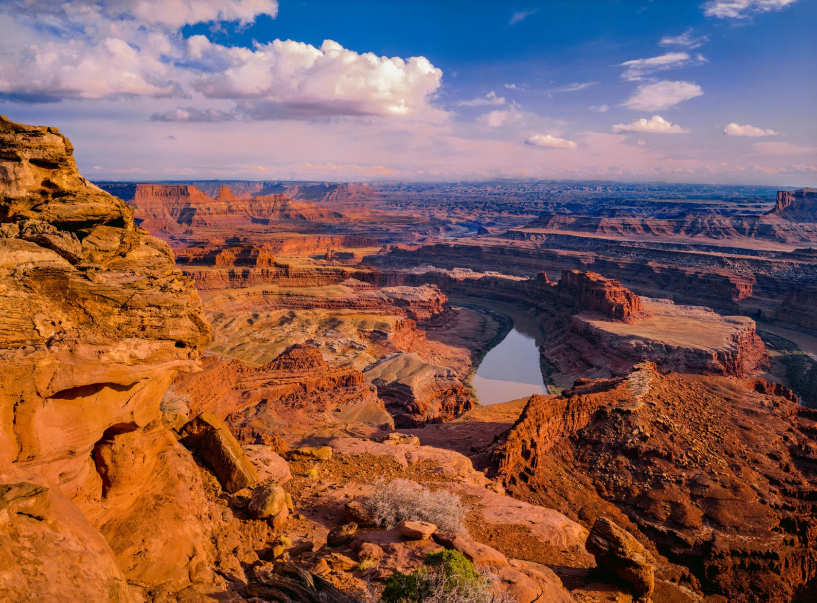 Aerial view of a vast canyon landscape with a river winding through red rock formations under a blue sky with clouds.
