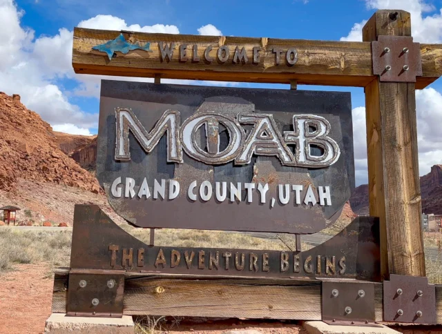 Entrance sign reading "Welcome to Moab, Grand County, Utah. The Adventure Begins" with desert landscape and rock formations in the background.