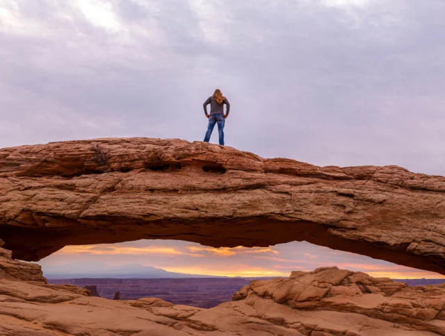 A person stands on a large, natural rock arch with a scenic view of a colorful sunset sky and distant landscape.
