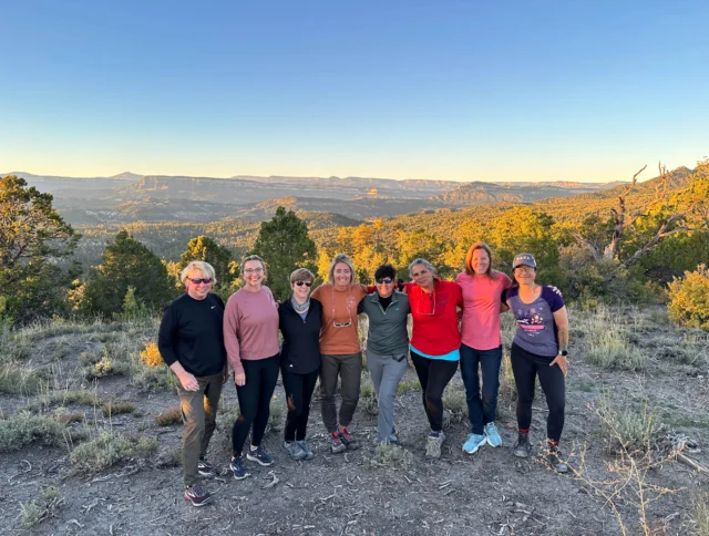 A group of eight people stands together on a grassy hill with a scenic mountain view in the background. The sky is clear and the weather appears sunny.