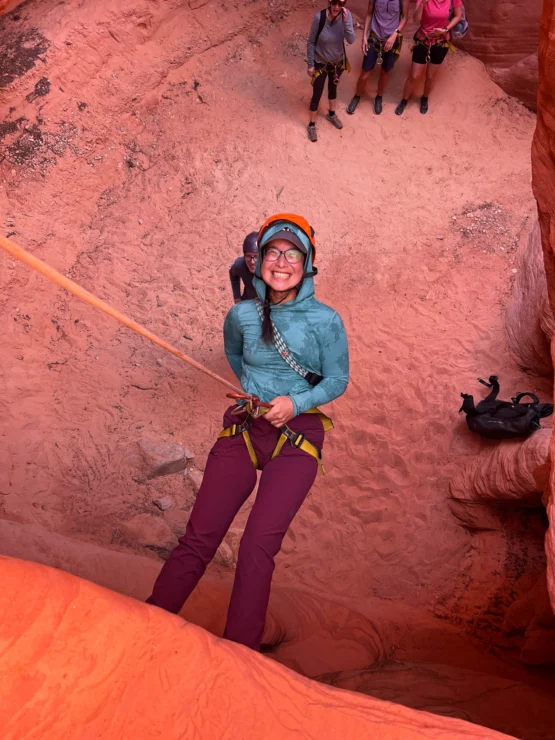 Person wearing climbing gear rappels down a red rock face, smiling. Three people stand at the top holding climbing ropes.