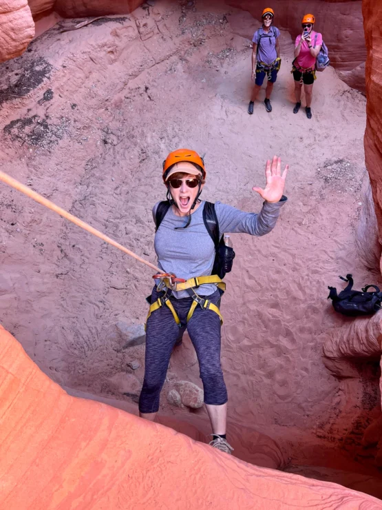 Person in climbing gear waves while rappelling down a rocky canyon, with two people in similar gear standing at the top.