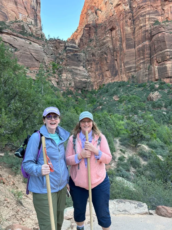 Two people with hiking sticks stand on a rocky trail surrounded by greenery and large canyon walls. They wear hats and sunglasses.