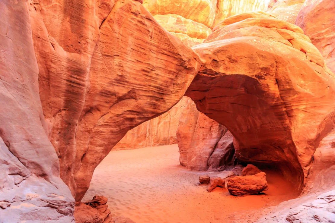 A natural sandstone arch in a desert landscape, with sunlight illuminating its red and orange hues, casting shadows on the sand below.