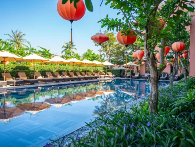 A serene poolside scene with lounge chairs, large red lanterns, and yellow umbrellas. Palm trees and greenery surround the area, reflecting in the water.