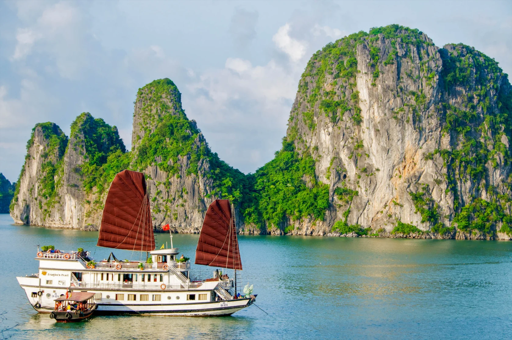 A white boat with red sails travels through clear waters, surrounded by large, rocky green cliffs under a blue sky.