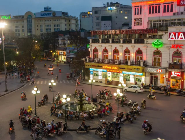 A bustling roundabout in an urban area at dusk, with numerous motorcycles and cars surrounding a central fountain, against a backdrop of illuminated buildings and storefronts.