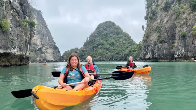 Three people kayaking on calm water between rocky cliffs and lush hills under a cloudy sky.