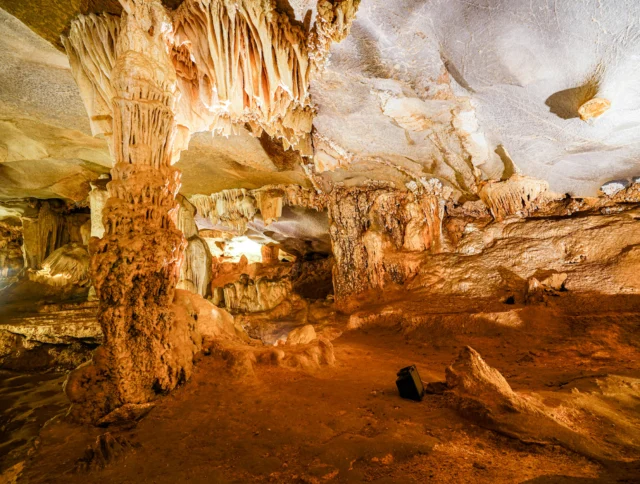 A spacious cave interior with jagged stalactites hanging from the ceiling and stalagmites rising from the ground, illuminated by soft, warm lighting.