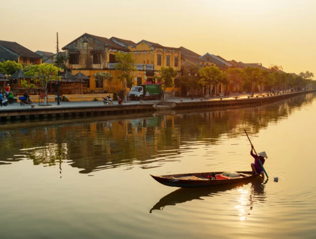 A person rows a boat on a calm river at sunset, with historic buildings and trees lining the riverbank in the background.