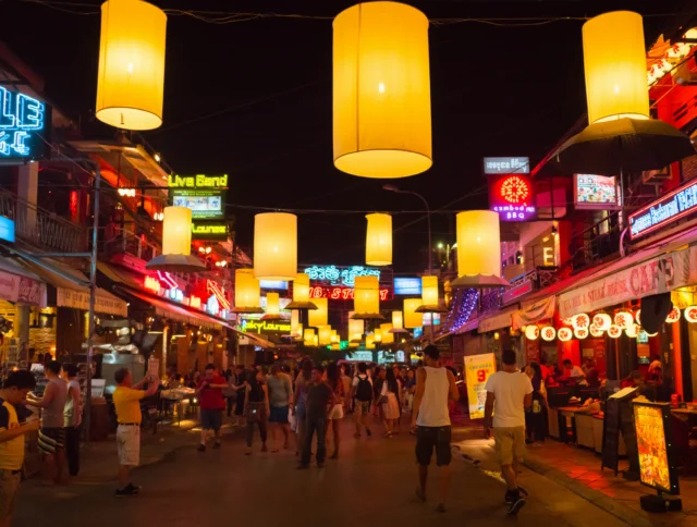 A lively street market at night with large hanging lanterns and neon signs. People are walking and socializing along the vibrant, illuminated street.