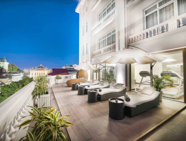 A rooftop terrace with lounge chairs, umbrellas, and greenery against a night sky, attached to a white building with large windows.