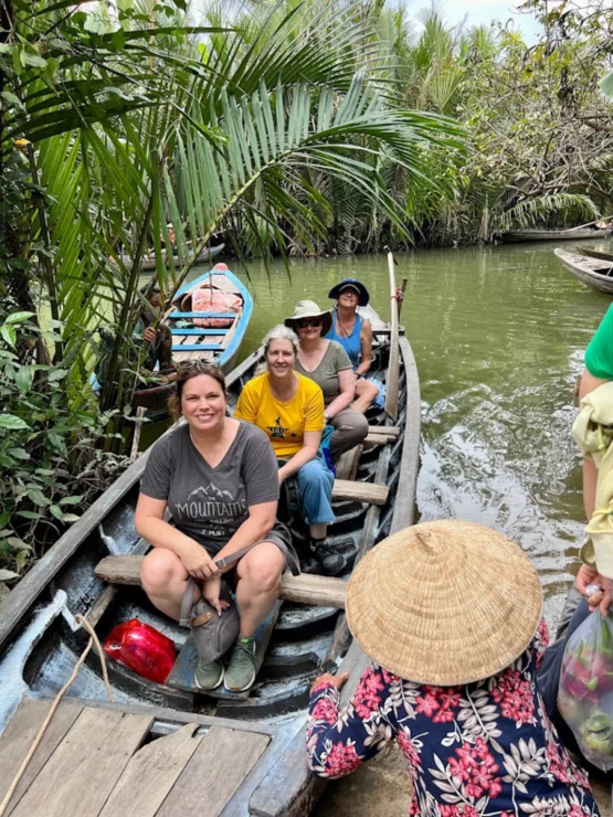 People seated in a wooden boat on a narrow tropical waterway, with lush greenery. A person in a conical hat paddles at the front.