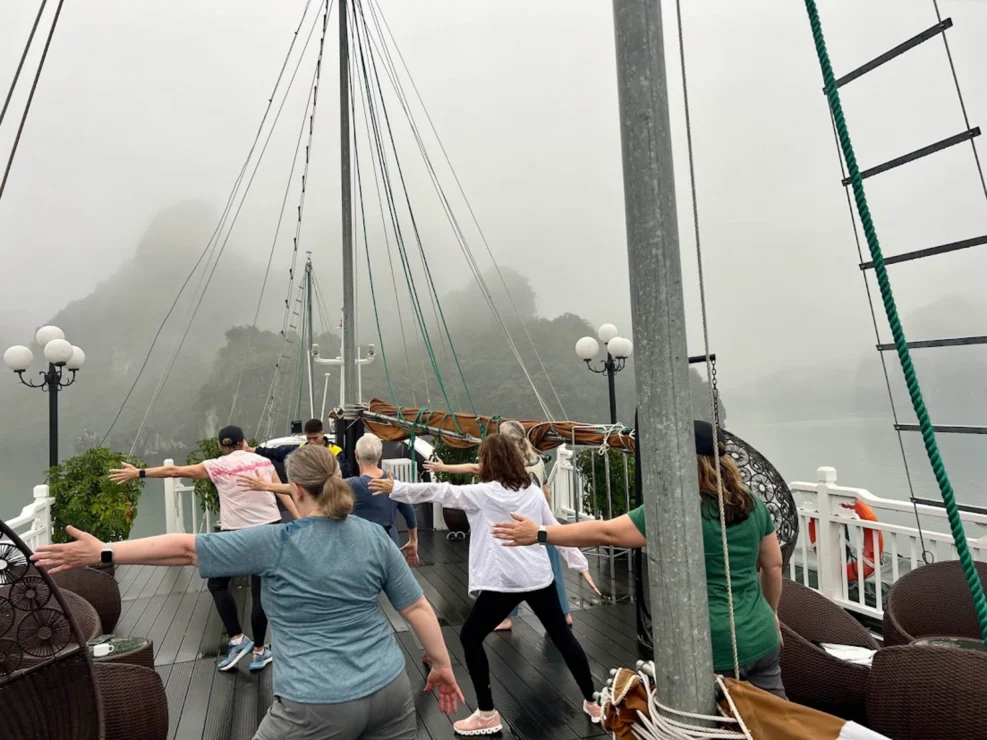 People practice yoga or tai chi on a boat deck with misty mountains in the background.