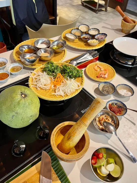 A cooking station with various ingredients, including herbs, spices, citrus, a large fruit, and mortar and pestle on a countertop.