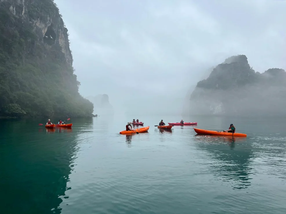 People kayaking on calm water surrounded by misty cliffs and greenery.