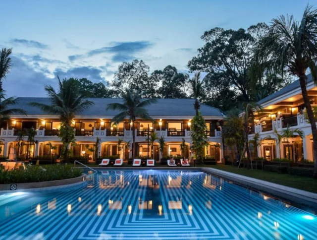 A hotel pool area at dusk with illuminated rectangular pool, surrounded by palm trees and two-story buildings with balconies.