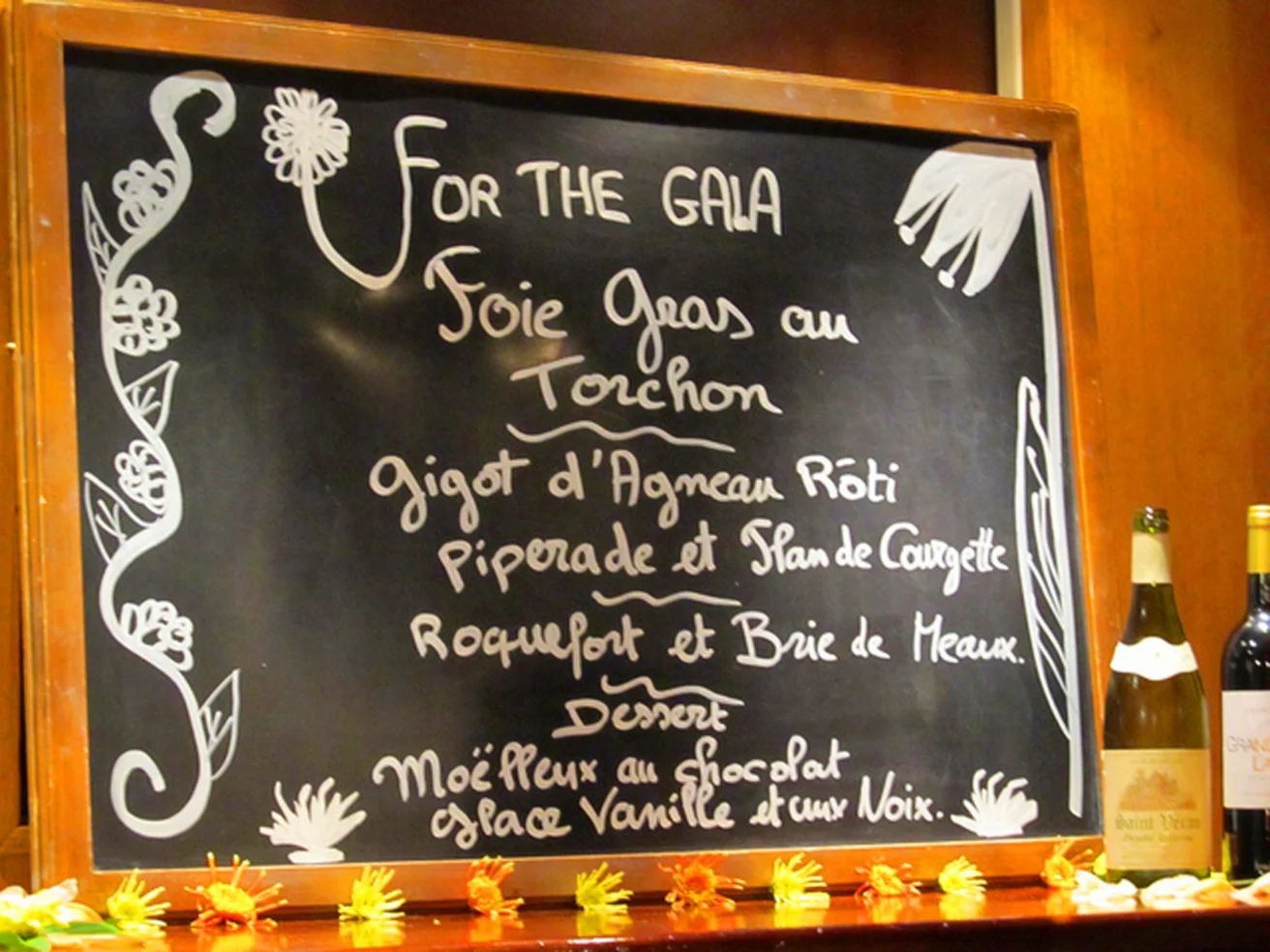 Chalkboard menu for a gala featuring foie gras, roasted lamb, piperade, Roquefort and Brie, and a chocolate dessert. Bottles of wine are placed nearby.