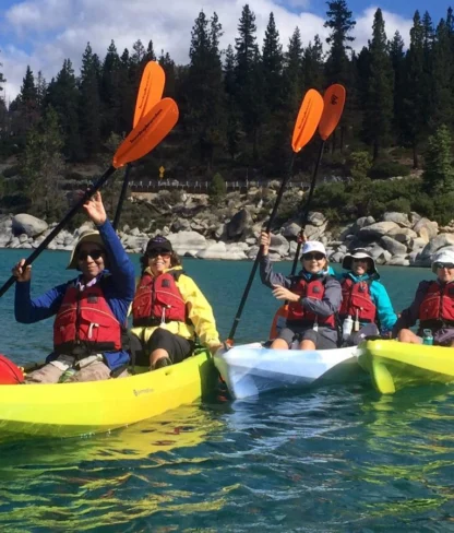 Group of five people kayaking on a clear lake surrounded by trees and rocks, holding orange paddles above their heads.
