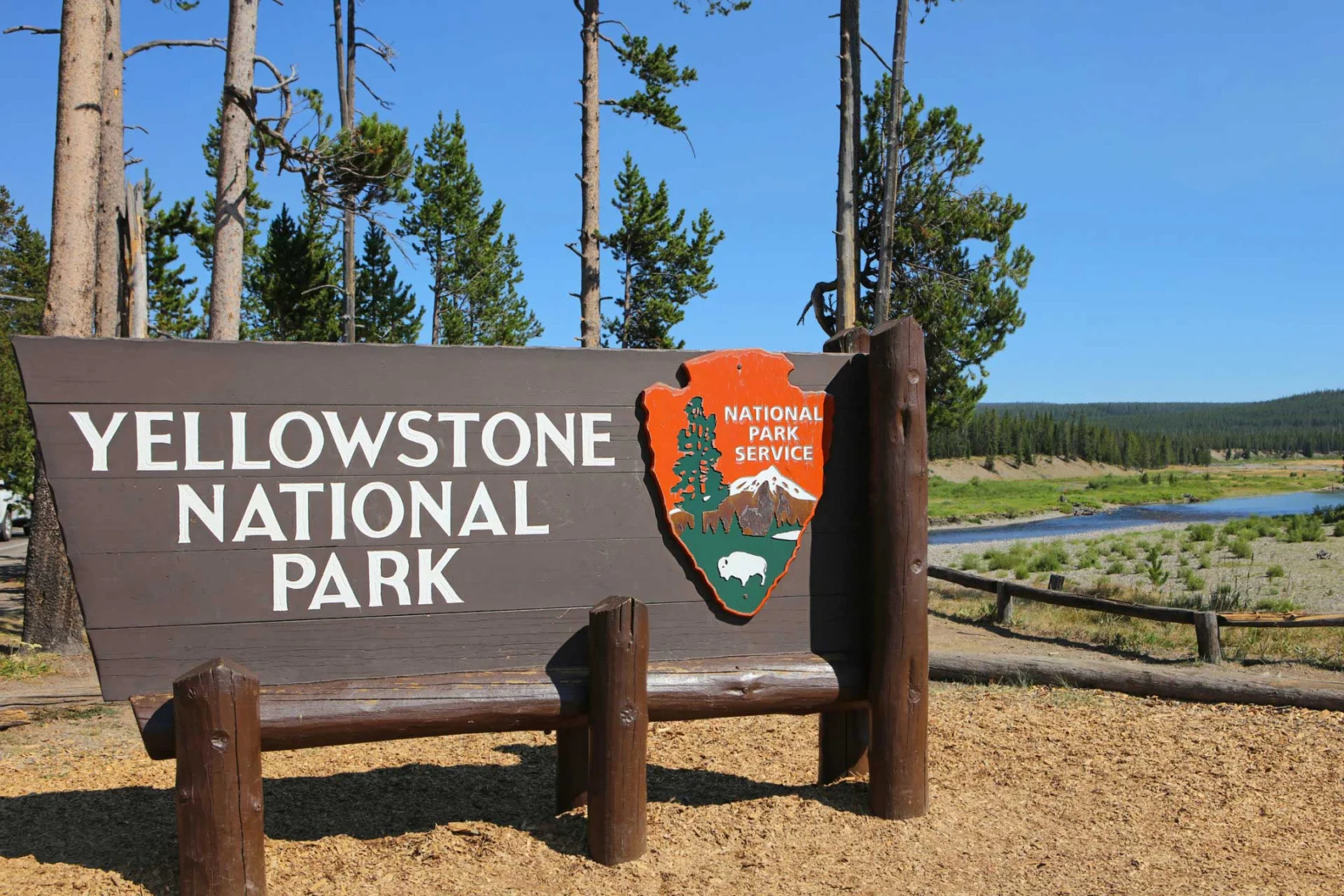 Sign for Yellowstone National Park with trees and a river in the background under a clear blue sky.