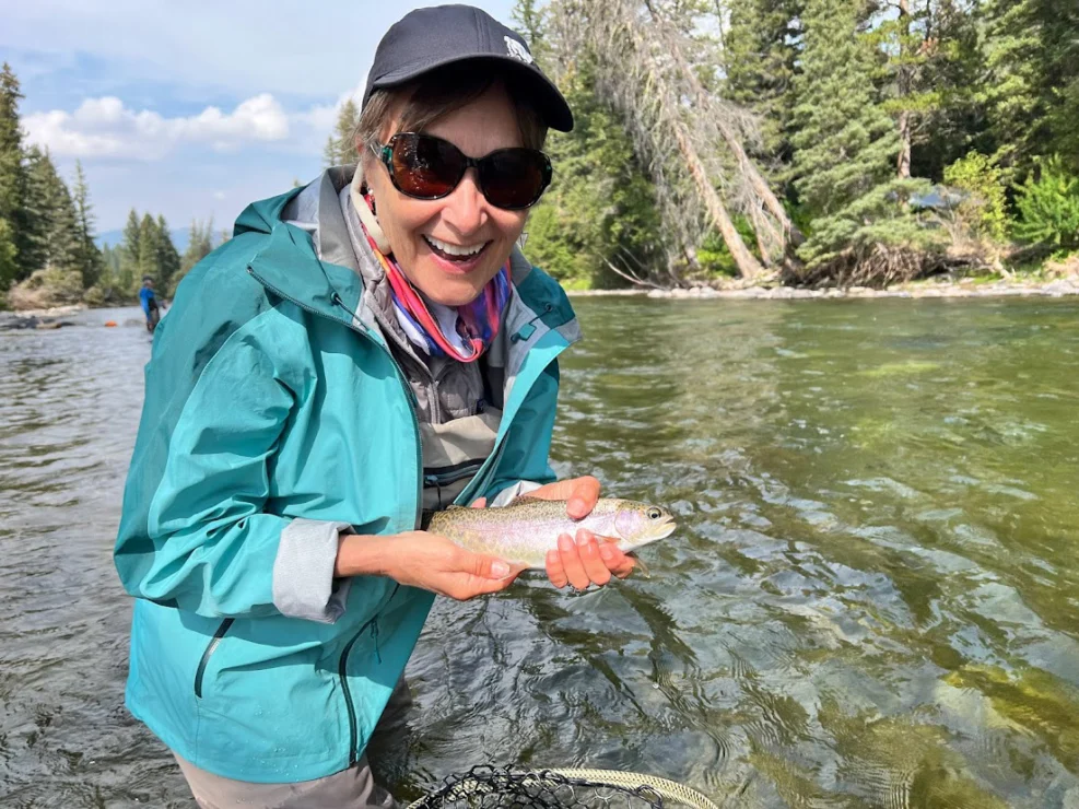 Person smiling, holding a fish in a river. Wearing a blue jacket, cap, and sunglasses. Trees in the background.