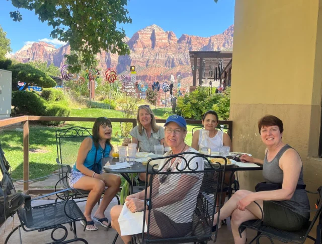 Five people sitting at an outdoor table, enjoying a meal with mountains visible in the background.