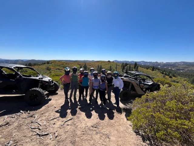 A group of people in helmets stand between two off-road vehicles on a dirt path with a scenic mountainous background.