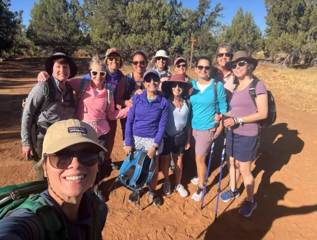 A group of eleven people wearing hiking gear stand together outdoors on a sunny day, with trees and a dirt path in the background.