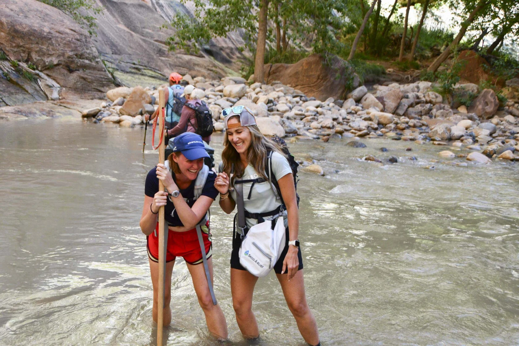 Two people smiling and wading through a rocky river with walking sticks. Others are visible in the background. They appear to be hiking in a natural setting with trees and large rocks.