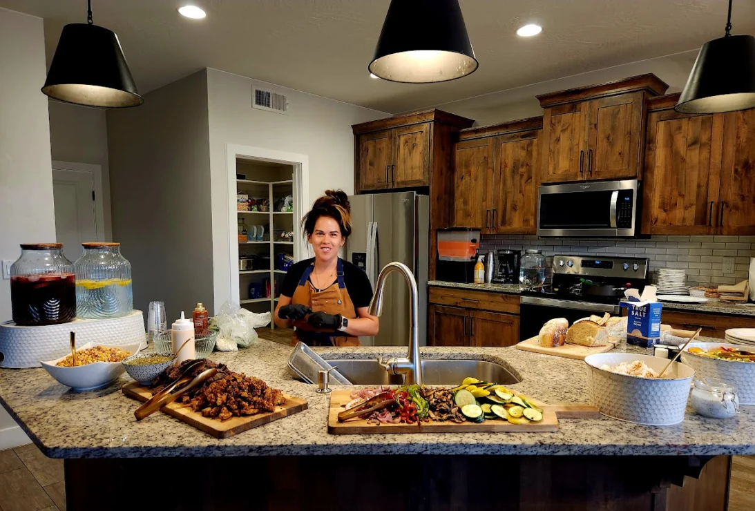 A woman stands in a kitchen, preparing food on a granite island with various dishes and ingredients. Wooden cabinets and stainless steel appliances are in the background.